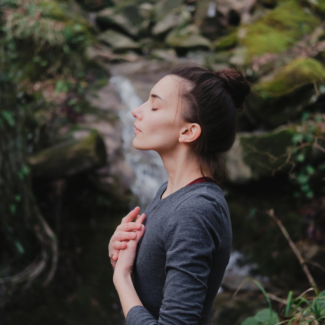 Young woman practicing breathing yoga pranayama outdoors in moss forest on background of waterfall. Unity with nature concept.